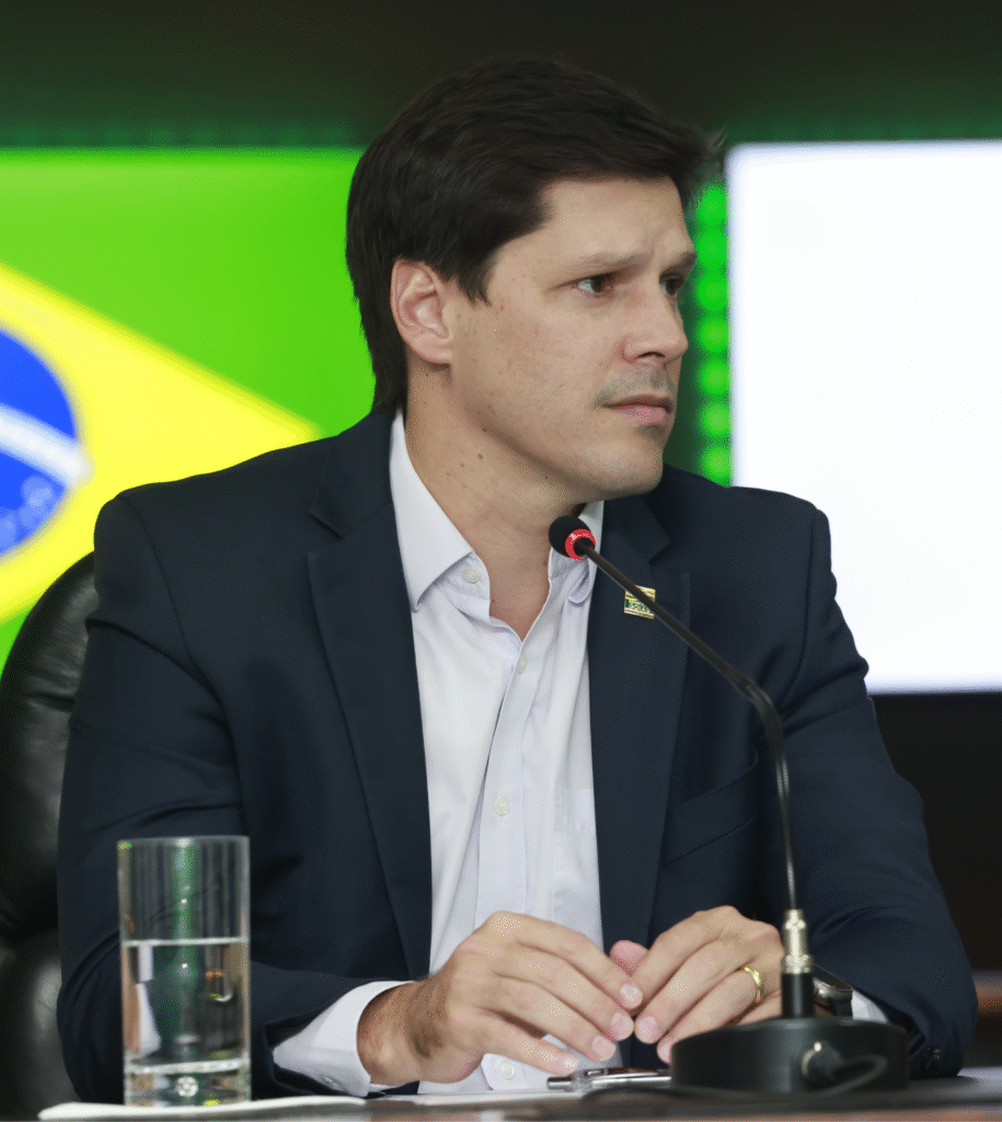 Daniel Vilela usando um terno. Está sentado com expressão concentrada. Participa de uma conferência. No fundo, pode-se ver a bandeira do Brasil em um painel de LED.