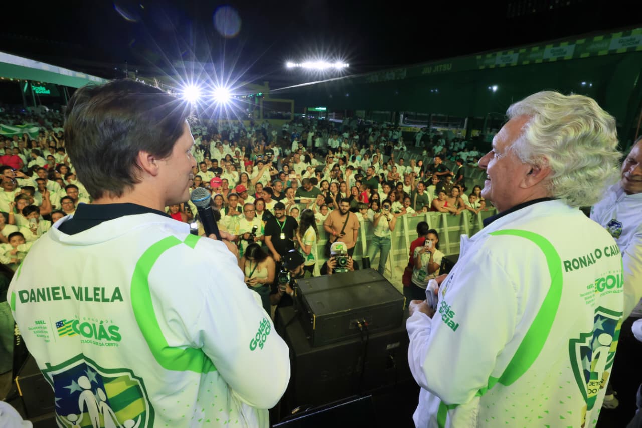 Daniel Vilela e Caiado de costas para a câmera. Ocupam um palco e falam para o público que participa do evento.