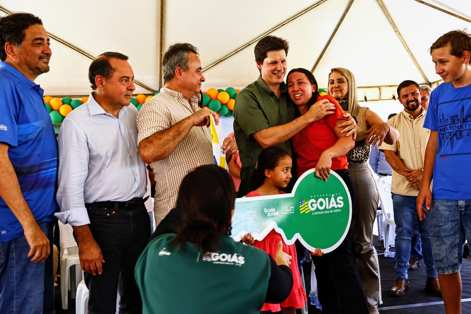 Na foto, Daniel Vilela está em cima de um palco junto com dois políticos que fazem parte do programa "Para Ter Onde Morar" da cidade de Jaupaci. Daniel está abraçando uma das beneficiadas do projeto; ela está segurando uma chave grande de papel que representa a conquista. Daniel Vilela está usando uma blusa verde e os demais estão de tons claros.