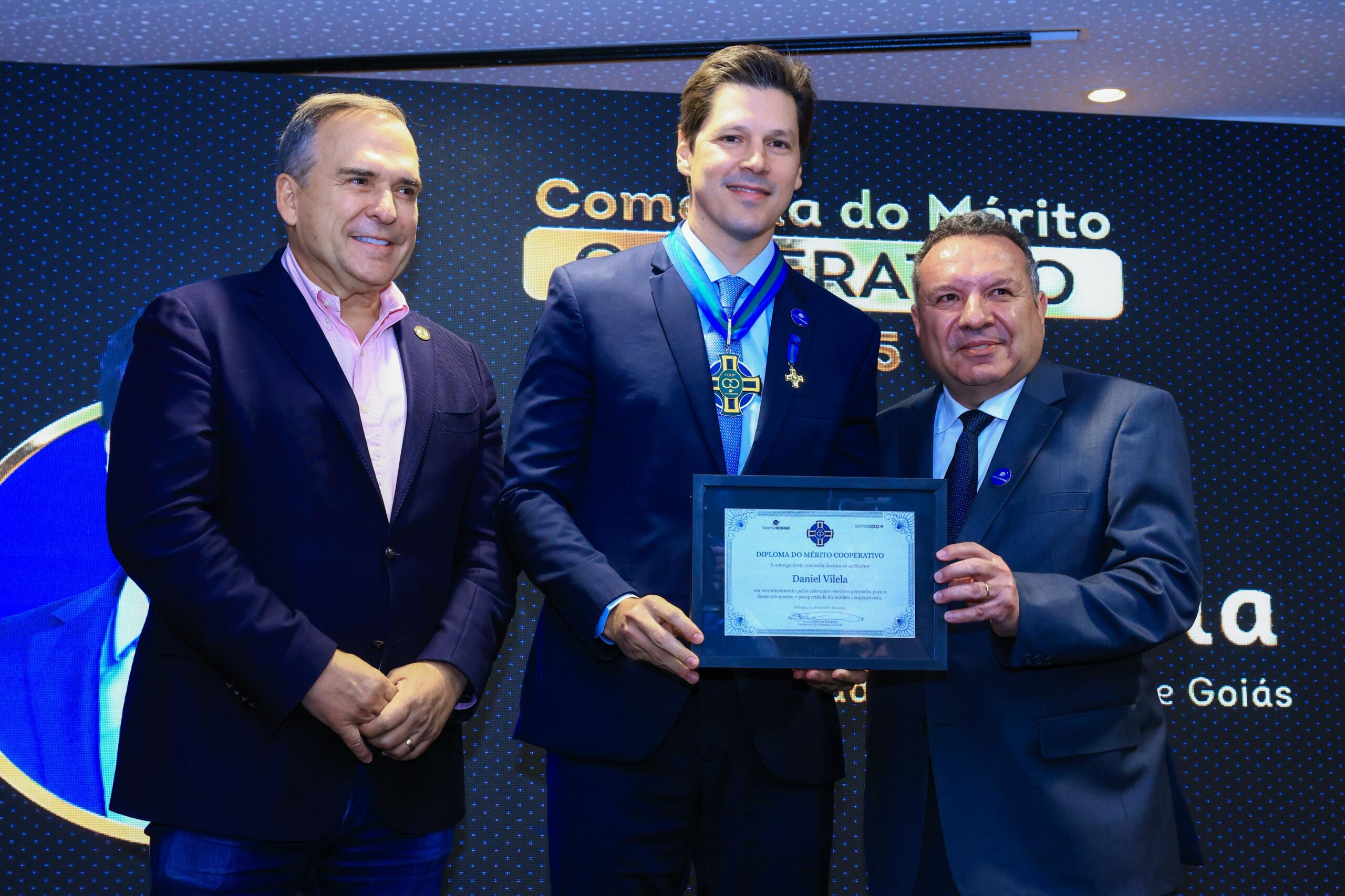 Em cima de um palco está Daniel Vilela, Sandro Mabel, prefeito de Goiânia, e o presidente do Sistema OCB/GO, Luís Alberto Pereira. Daniel está recebendo o diploma do Mérito Cooperativo, da OCB/GO. Os três usam terno e estão sorrindo para a foto. 